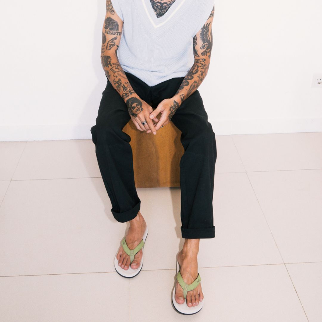 Man with tattoos on arms wearing white vest, black trousers and white and green mens flip flops