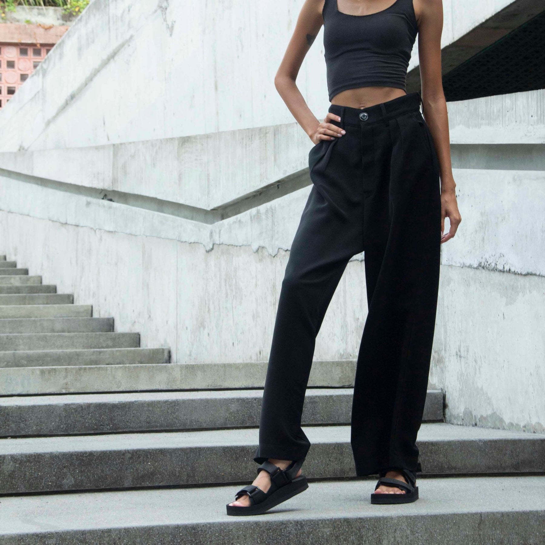 Woman wearing black trousers and black crop top and black adventure sandals standing on concrete steps